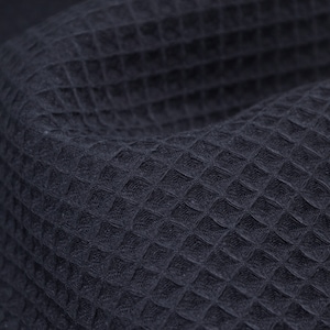 May include: Close-up of a navy blue waffle-weave fabric. The fabric has a textured surface with a grid-like pattern of small, raised squares. The fabric is slightly curved, showing the texture and depth of the material.