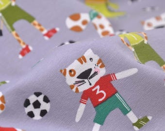 May include: Gray fabric with a repeating pattern of cartoon animals playing soccer. The pattern includes a cat wearing a red and green soccer uniform with the number 3 on the front, a giraffe wearing a green and yellow uniform, and a soccer ball.