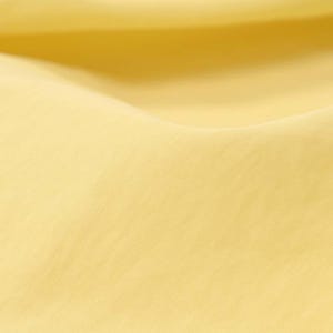 Puede incluir: Primer plano de una tela amarilla suave con textura sutil. El material está suavemente drapeado, creando curvas y sombras fluidas. La impresión general es de un textil liso y ligero.