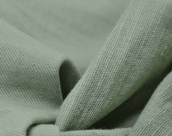 Könnte beinhalten: Nahaufnahme eines weichen, salbeigrünen Stoffes mit einer strukturierten Webart. Der Stoff ist gefaltet und drapiert, wodurch die Textur und der Fall des Materials sichtbar werden.