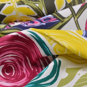 Può includere: Primo piano di un tessuto con un vivace motivo floreale. Il design presenta grandi fiori e foglie stilizzate nei toni del giallo, rosa, verde e blu su uno sfondo bianco. Il tessuto è piegato, mostrando la consistenza e la profondità del materiale.