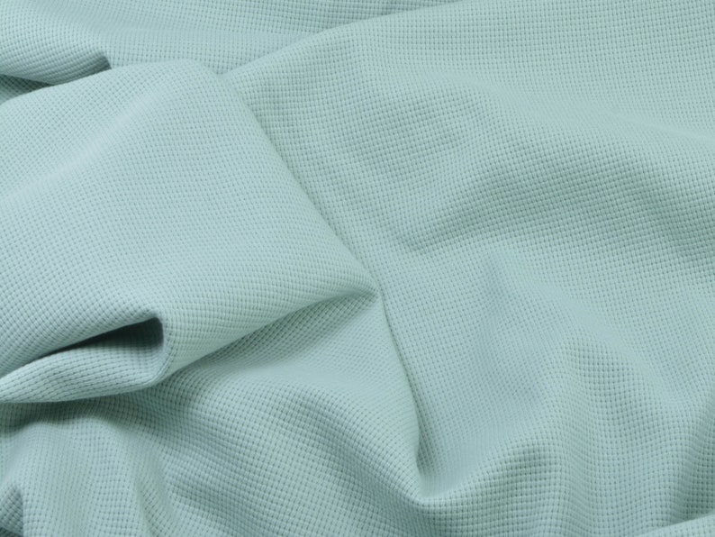 May include: A close-up of a light blue waffle knit fabric. The fabric is soft and textured, with a subtle sheen.