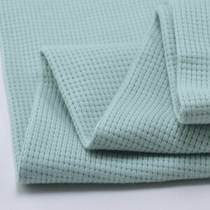 May include: A close-up of a light blue waffle knit fabric. The fabric is folded in half and the texture of the knit is visible.