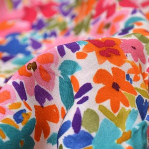 Op de afbeelding: Witte stof met een kleurrijke bloemenprint. De bloemen zijn abstract en geschilderd in tinten roze, oranje, blauw, paars en groen.