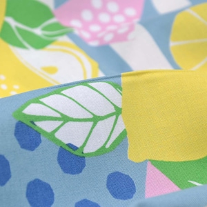 May include: Close-up of a fabric with a repeating pattern of lemons, leaves, and polka dots. The design features yellow lemons, green leaves, pink shapes, and blue polka dots on a light blue background. The fabric is folded.