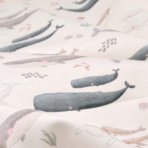 Peut inclure: Un tissu blanc avec un motif répété de baleines, de coraux et d'algues. Les baleines sont dans différentes nuances de gris et de rose.