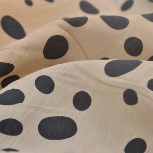 Op de afbeelding: Close-up van een beige stof met grote, donkergrijze stippen. De stof is gevouwen, waardoor zachte rondingen en schaduwen ontstaan. De stippen variëren in grootte en vorm, wat de textiel visueel aantrekkelijk maakt.