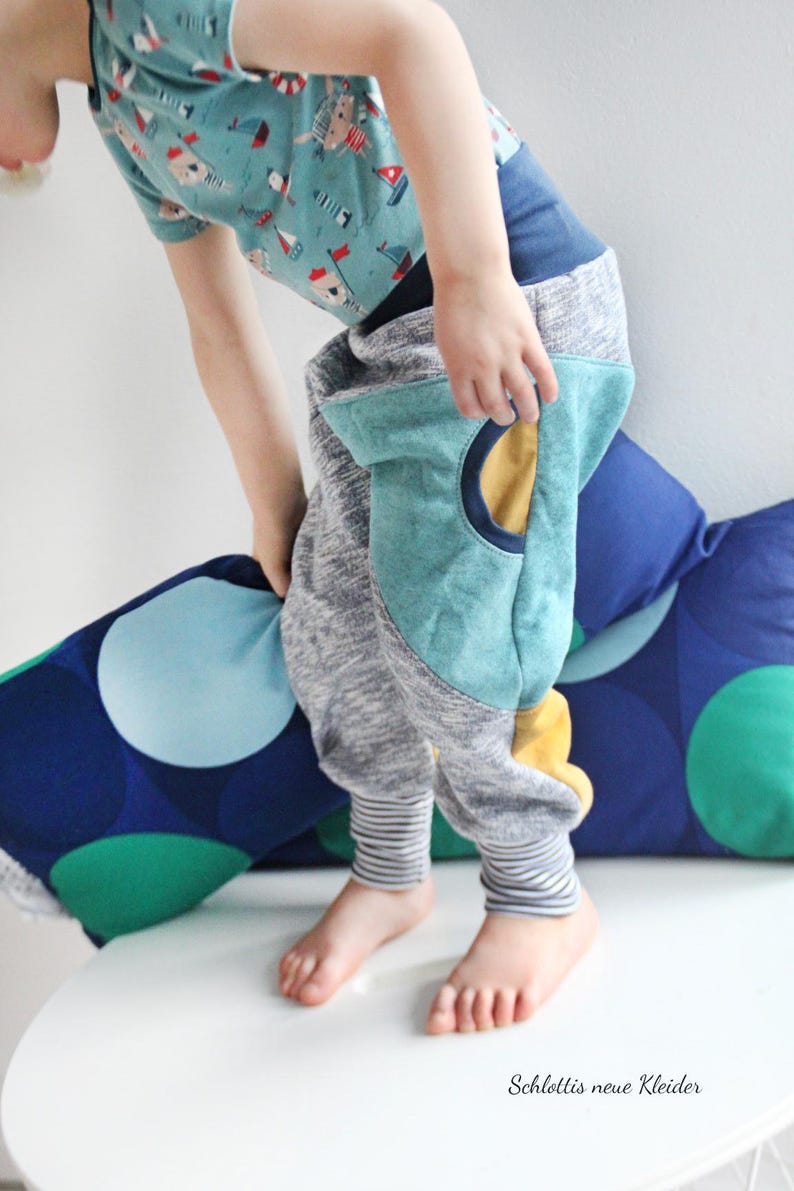 May include: A child's outfit featuring gray sweatpants with teal knee patches and striped cuffs. The outfit includes a light blue t-shirt with a nautical print. The text "Schlottis neue Kleider" is visible.
