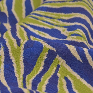 Pode incluir: Close-up de um tecido com um padrão de listras de zebra azul, verde e branco sujo. O tecido está dobrado, mostrando a textura e as variações de cor. O design apresenta listras arrojadas e irregulares, criando um efeito visual dinâmico. O tecido parece leve.