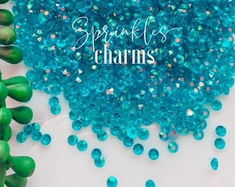 May include: A close-up image of turquoise blue iridescent glass rhinestones. The rhinestones are scattered on a white surface. The text "Sprinkles charms" is visible in the image.
