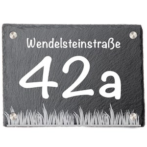 Könnte beinhalten: Rechteckiges, dunkelgraues Schiefer-Hausnummernschild mit dem weißen Text "Wendelsteinstraße 42a". Das Schild hat unten ein dekoratives Grasdesign und ist mit vier silbernen Nieten befestigt. Der Hintergrund ist weiß.