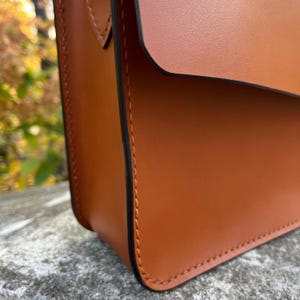 May include: Close-up of a brown leather bag with a flap closure and black stitching. The bag has a smooth, textured surface.