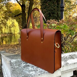 May include: A brown leather briefcase with a top handle and a gold metal clasp. The briefcase is sitting on a stone surface.