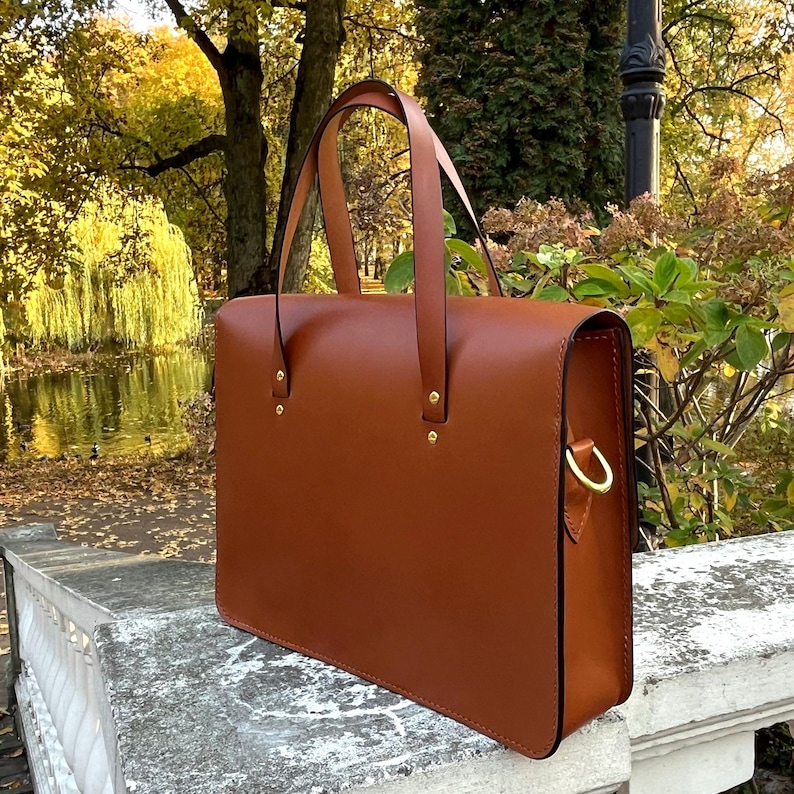May include: A brown leather briefcase with a top handle and a gold metal clasp. The briefcase is sitting on a stone surface.