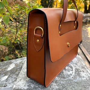 May include: A brown leather satchel bag with a top handle and a shoulder strap. The bag has a flap closure with a brass buckle.