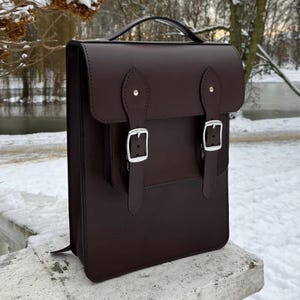 Puede incluir: Una mochila tipo cartera de cuero marrón oscuro con asa superior y dos cierres de hebilla. La bolsa tiene un bolsillo frontal y herrajes plateados. La mochila está sobre una superficie de piedra con un fondo nevado.