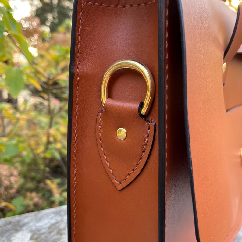 May include: Close-up of a brown leather satchel bag with a gold metal ring and a leather flap. The bag has a black stitched edge.