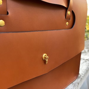May include: A brown leather satchel bag with a gold metal clasp and decorative hardware. The bag has a flap closure and a top handle.