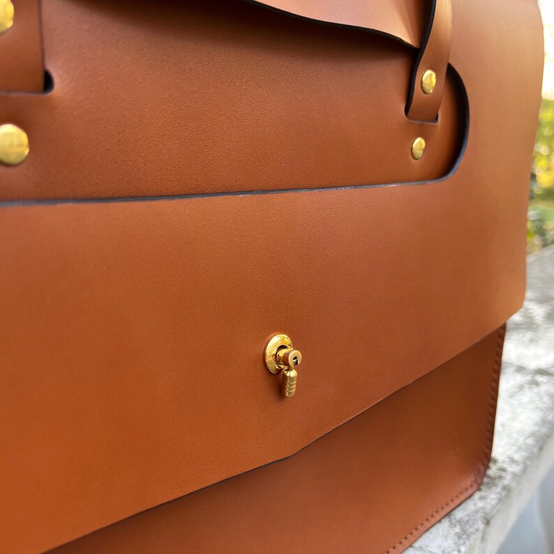 May include: A brown leather satchel bag with a gold metal clasp and decorative hardware. The bag has a flap closure and a top handle.