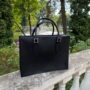 May include: A black leather tote bag with two top handles and gold-coloured hardware. The bag has a rectangular shape with a structured design. The background features a park setting with trees and a fountain.