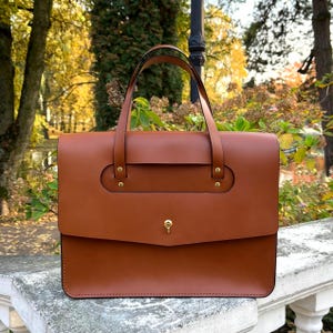 May include: A brown leather briefcase with two top handles and a gold clasp. The briefcase has a flap with a small pocket and a gold button.