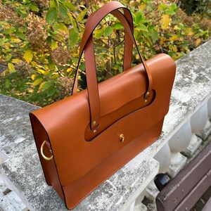 May include: A brown leather briefcase with a top flap and a single strap. The briefcase has gold hardware and a decorative detail on the front flap.