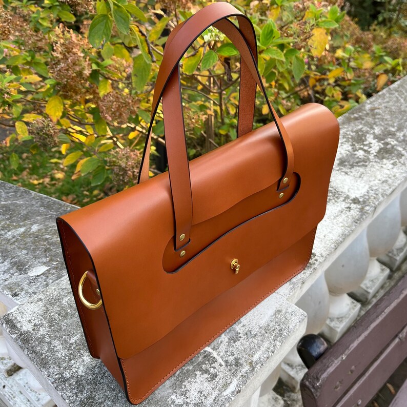 May include: A brown leather briefcase with a top flap and a single strap. The briefcase has gold hardware and a decorative detail on the front flap.