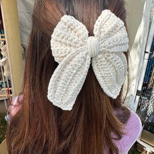 May include: A large, cream-colored crochet bow hair clip. The bow is made of thick yarn, with a textured, ribbed pattern. The bow is centered and attached to a clip, designed to be worn in the hair. The hair accessory is a handmade item.