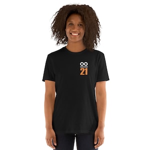 Op de afbeelding: Een zwart t-shirt met een kleine afbeelding op de linkerbovenborst. De afbeelding bevat het oneindigheidssymbool en het nummer 21 in wit en oranje. Het shirt heeft een ronde hals en wordt door een persoon gedragen.
