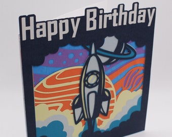 May include: A blue card with the words "Happy Birthday" in white lettering. The card features a paper cut-out design of a rocket ship flying through space with planets and stars.