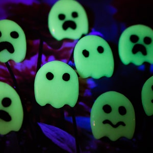 Op de afbeelding: Een groep van acht glow-in-the-dark ornamenten in de vorm van een geest met zwarte gezichtskenmerken. De geesten zijn gerangschikt op een paarse en zwarte achtergrond.