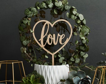 May include: A wooden heart-shaped sign with the word "love" written in cursive, placed in a white ceramic pot with a green leafy plant. The pot is surrounded by other green plants and two gold metal candle holders.