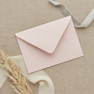 May include: A light pink envelope on a beige fabric background with a white ribbon and a light gray ribbon.