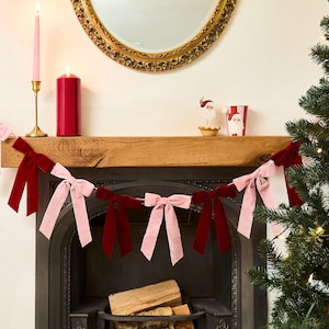 Pode incluir: Uma lareira festiva decorada para as festas. Uma guirlanda de laços de veludo rosa e bordô está pendurada em frente à lareira. Uma vela rosa acesa está em um castiçal dourado, e uma vela vermelha está ao lado.