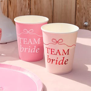May include: Two pink paper cups with the text "TEAM bride" printed on them. The cups have a white interior and a decorative bow design. One cup is a darker pink, and the other is a lighter pink.