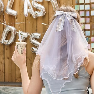 Peut inclure: Un voile de mariée blanc avec un nœud en sequins argentés. Le voile est porté par une personne aux cheveux longs blonds. Le voile est un accessoire populaire pour les enterrements de vie de jeune fille et les fêtes de mariage.