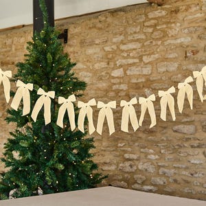 May include: A decorative garland featuring a series of uniform, cream-coloured fabric bows strung together in a horizontal line. Each bow has two long, hanging tails and is evenly spaced along the length of the string, creating a repeating pattern.