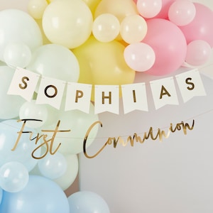 May include: A white banner with the name "Sophia" in gold letters, and a gold banner with the text "First Communion". The banners are hanging in front of a wall of pastel colored balloons.