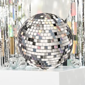 Peut inclure: Une grosse boule disco argentée avec des carreaux miroir. La boule est posée sur une surface blanche avec de plus petites boules disco en arrière-plan.