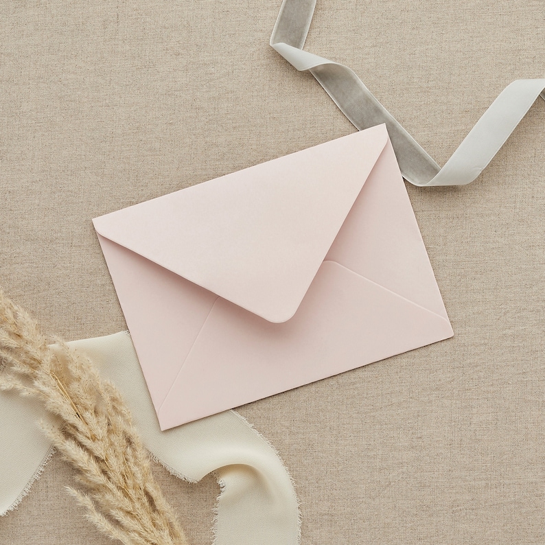 May include: A light pink envelope on a beige fabric background with a white ribbon and a light gray ribbon.
