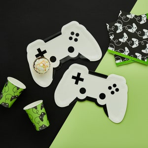 May include: Two white paper plates shaped like video game controllers with black details. The plates are on a black and green background. There are two small paper cups with a black and green design and a cupcake on one of the plates.
