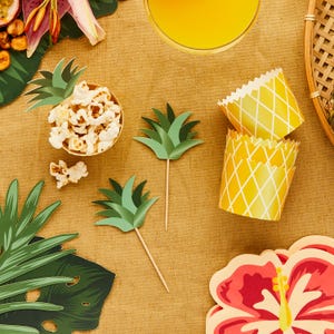 May include: A tropical party table setting with a yellow pineapple-themed cupcake liner, green paper pineapple toppers, a bowl of popcorn, and a red hibiscus flower.