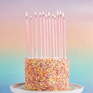 Może przedstawiać: Różowo-biały tort z różowymi świeczkami na wierzchu. Tort jest pokryty kolorowymi posypkami.