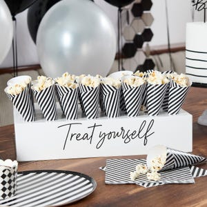 Puede incluir: Una caja blanca con conos de papel rayados en blanco y negro llenos de palomitas de maíz. La caja tiene el texto "Treat yourself" escrito en ella.