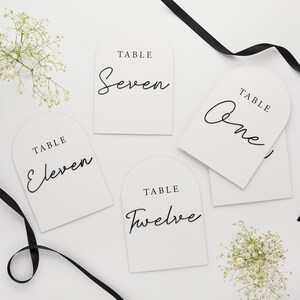 Può includere: Quattro carte di numero di tavolo bianche con calligrafia nera. Le carte hanno la forma di archi e hanno le parole "Tavolo" e i numeri di tavolo "Uno", "Sette", "Undici" e "Dodici" scritti su di esse.