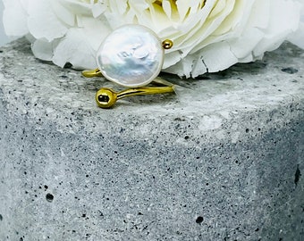 May include: A gold ring featuring a large, white pearl. The ring is displayed on a grey, textured surface.