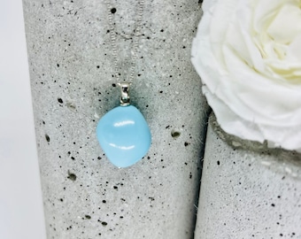 May include: A light blue gemstone pendant necklace on a silver chain. The pendant is a smooth, rounded shape and is hanging from a concrete surface.