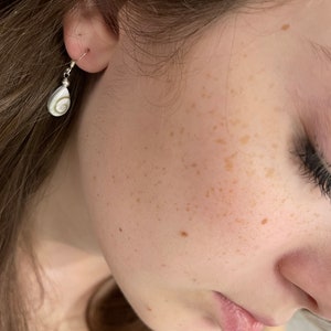 Peut inclure: Gros plan sur l'oreille d'une personne avec une boucle d'oreille en argent ornée d'un pendentif en coquillage blanc.