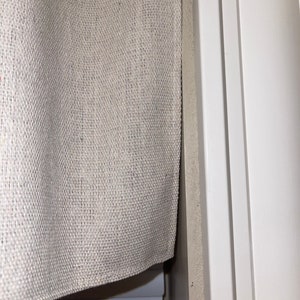 May include: A close-up of a beige burlap valance with a white window blind underneath. The valance is hanging on a white window frame.