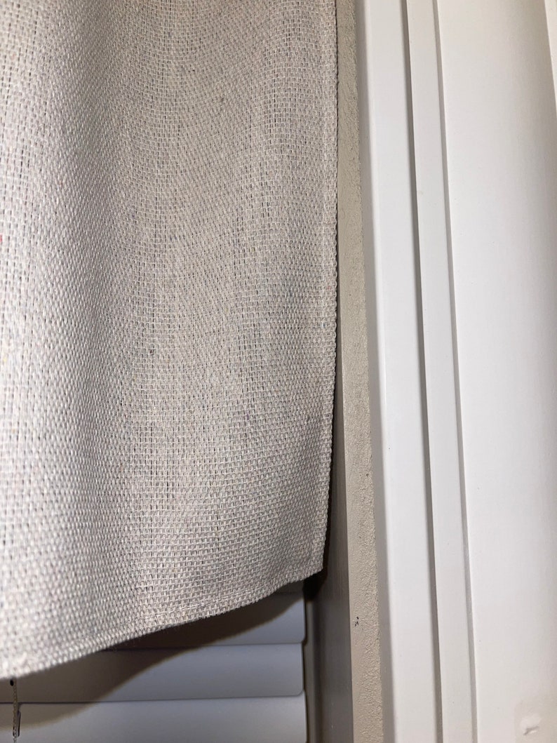 May include: A close-up of a beige burlap valance with a white window blind underneath. The valance is hanging on a white window frame.